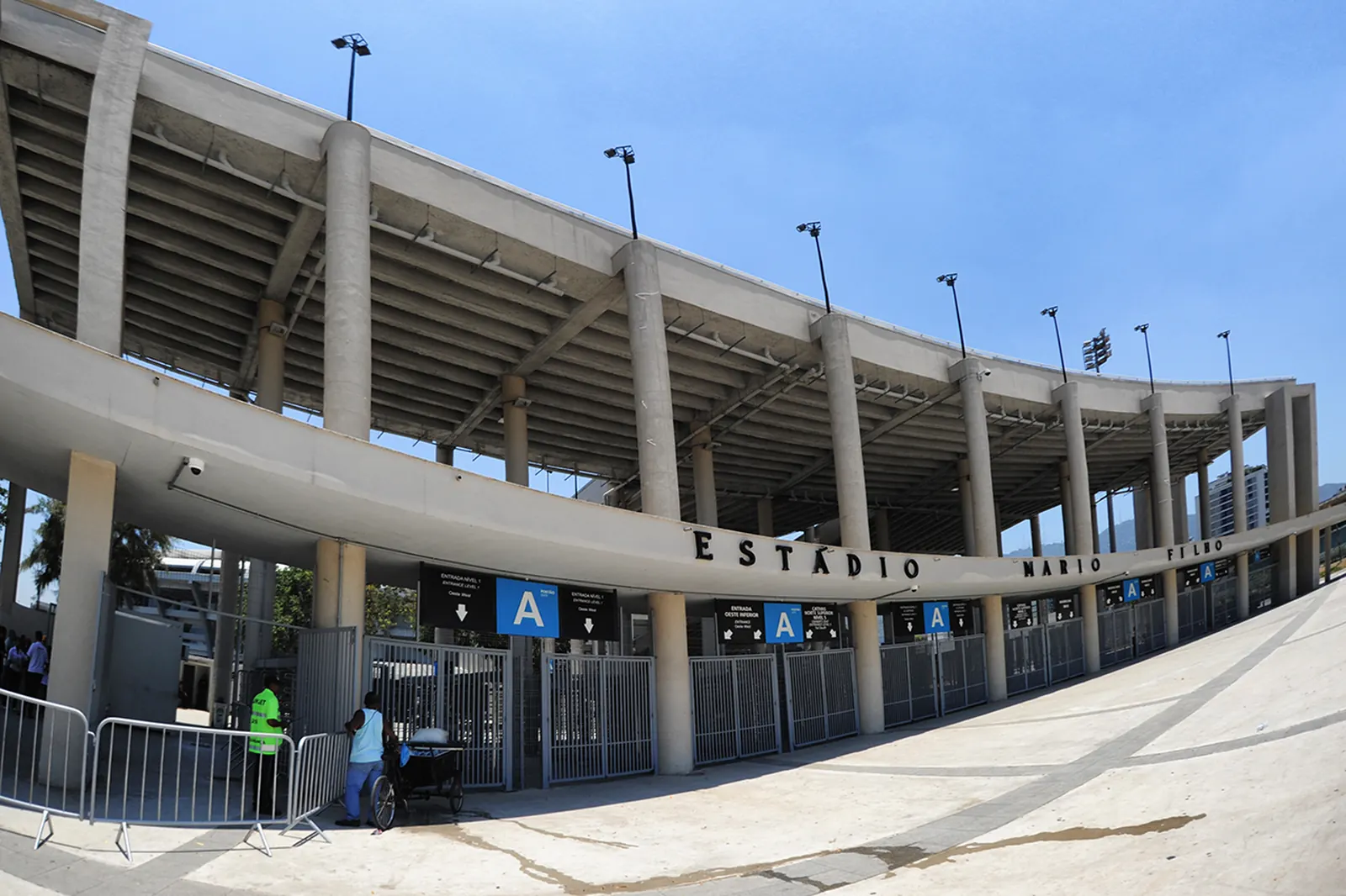 La facciata curva in cemento dello stadio in una giornata di sole, con un'insegna blu dell'ingresso contrassegnata dalla lettera A e palme lungo la piazza