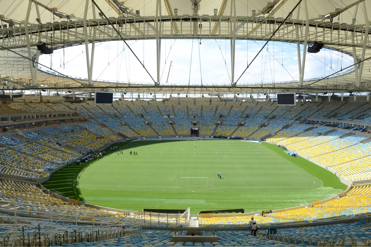 O interior do estádio do Maracanã, com bancadas de lugares amarelos, azuis e brancos em torno de um relvado verde sob uma cobertura em forma de anel