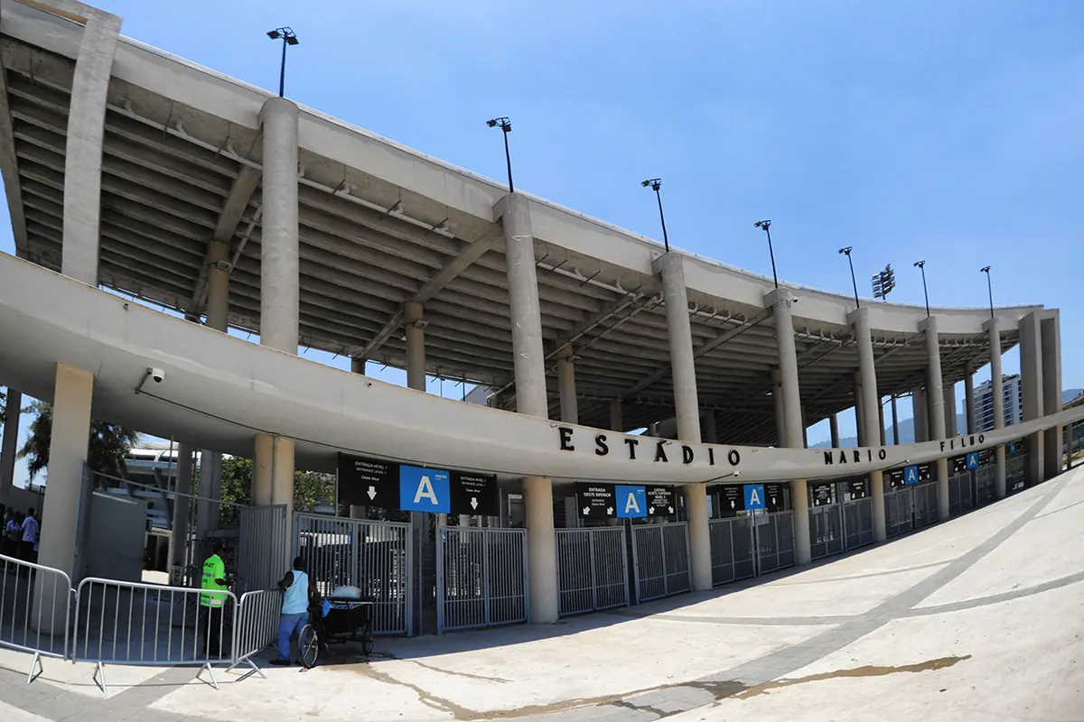 Zakřivené betonové průčelí Maracanã v slunečný den, s modrým vstupním označením A a palmami podél náměstí