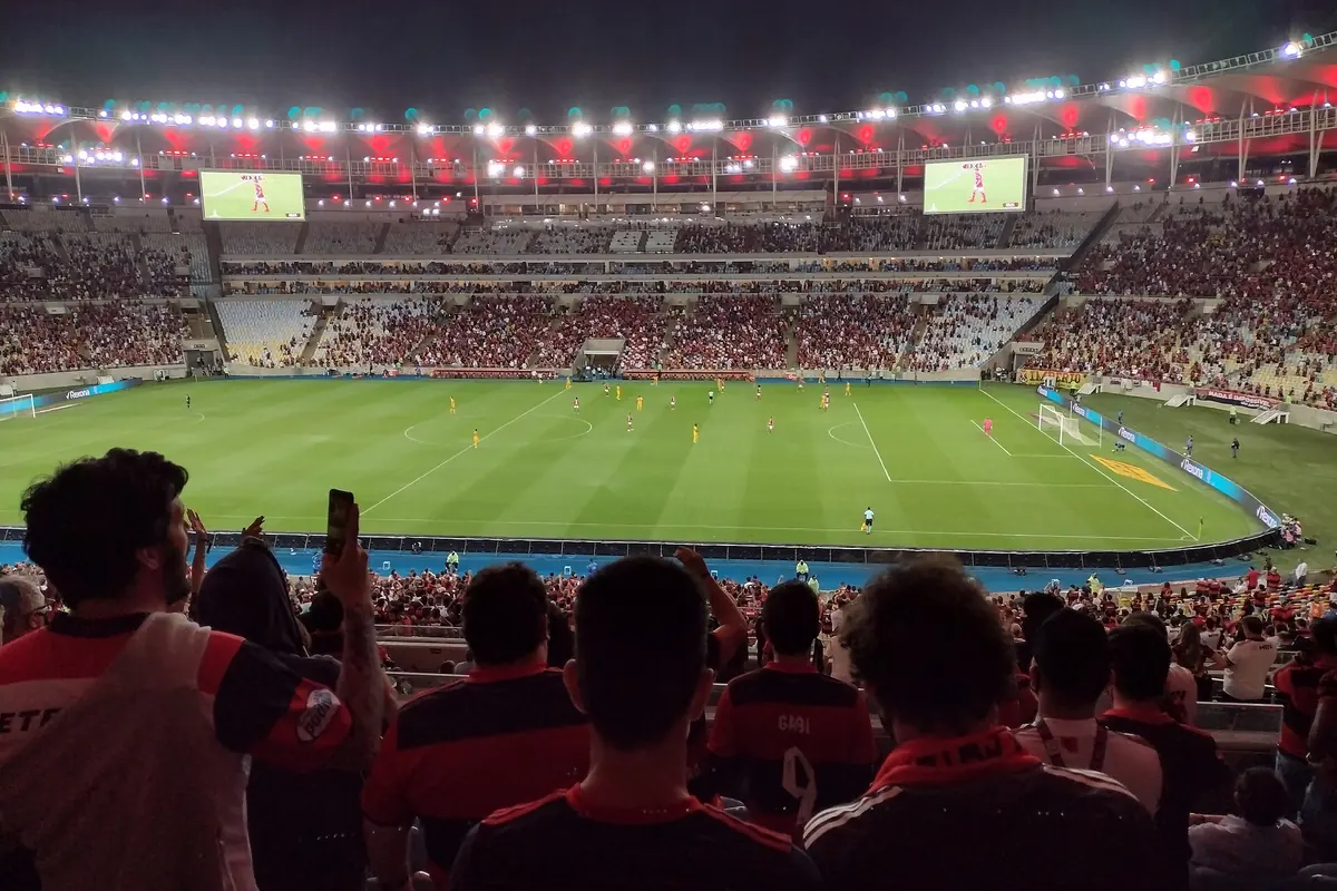 Um jogo no Maracanã com as bancadas cheias de adeptos de vermelho e preto sob a cobertura, visto do piso superior