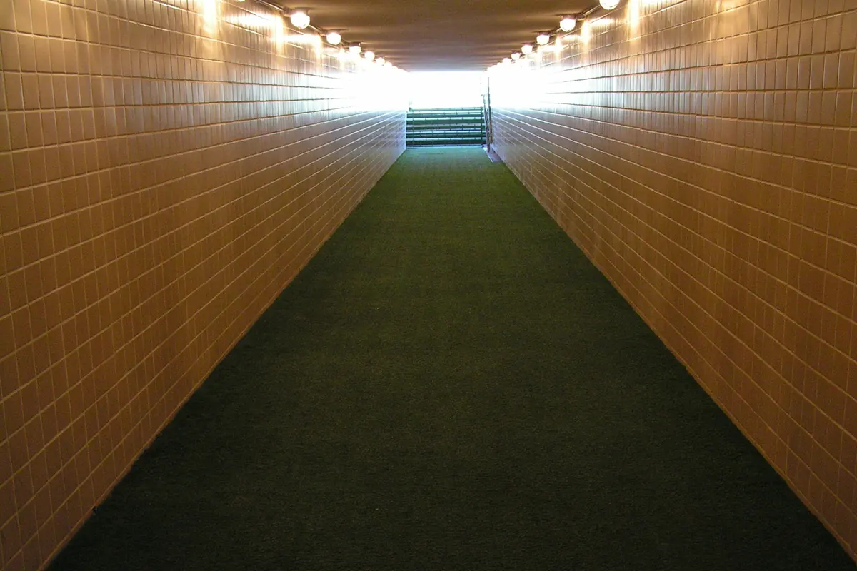 Um longo túnel de paredes castanho-alaranjadas e chão de alcatifa verde, iluminado por lâmpadas de teto, que conduz em direção à luz do dia