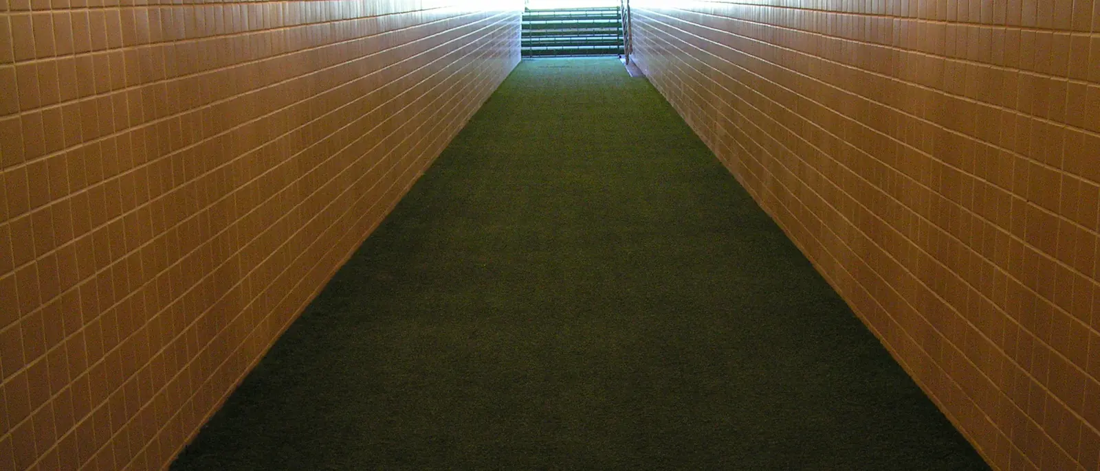 Un lungo tunnel con pareti color arancio-marrone e pavimento in moquette verde, illuminato da lampade a soffitto, che conduce verso la luce del giorno e un campo da calcio